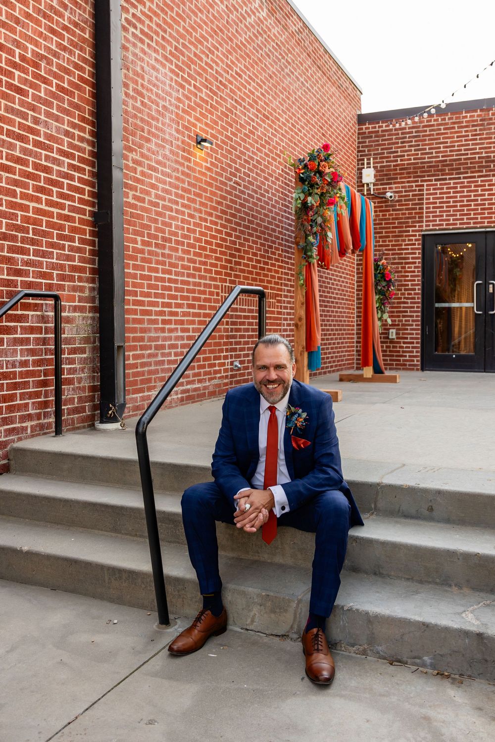 Smiling groom in navy suit and red tie sits on brick building steps with colorful floral decorations at Founders One Nine in Omaha, NE.