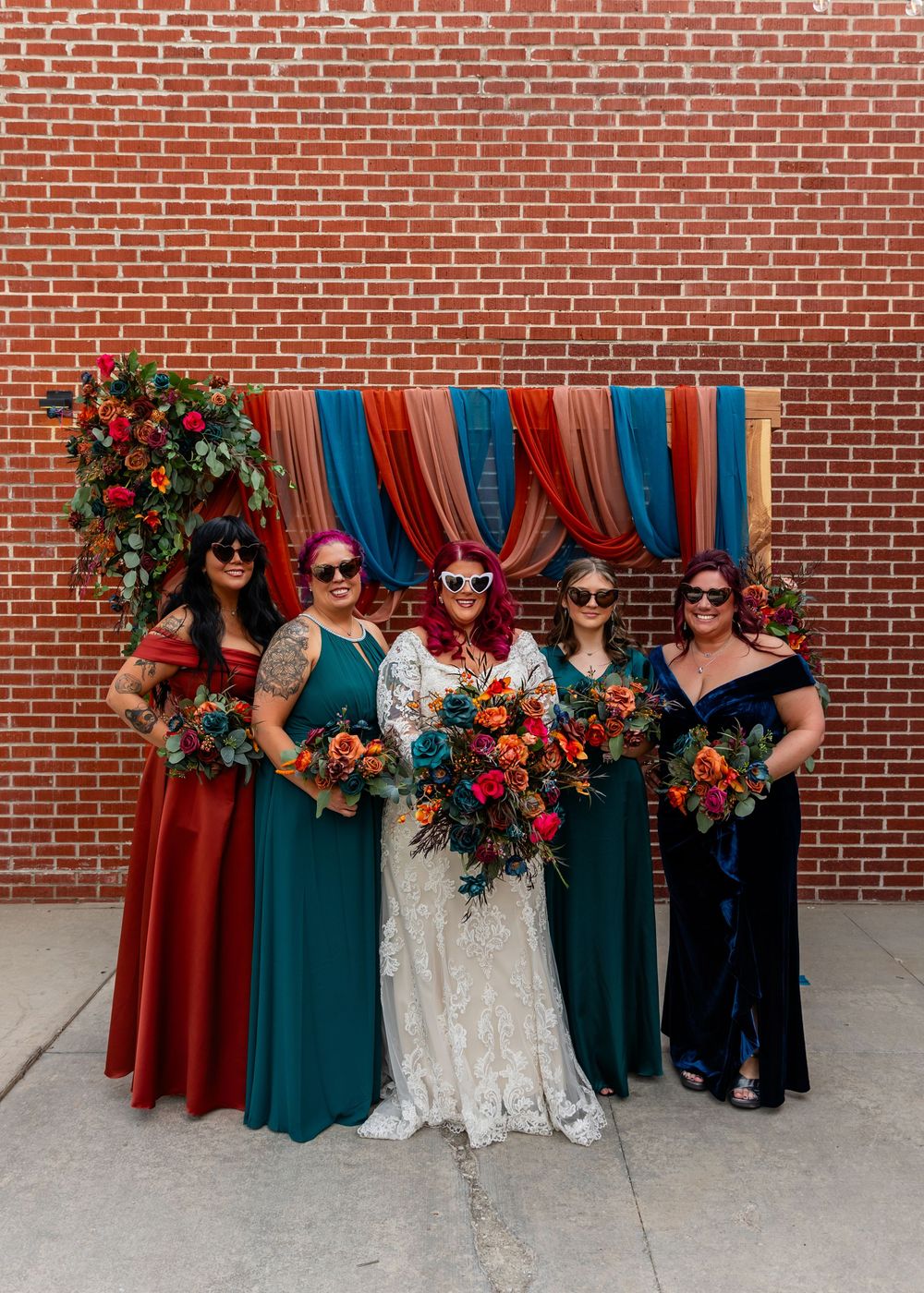 Bride in lace gown with four bridesmaids in colorful dresses holding vibrant bouquets before brick wall backdrop at Founders One Nine