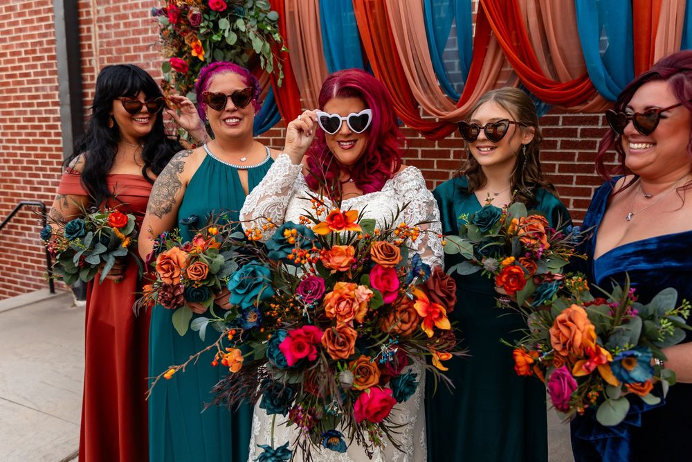 Bride in white dress with heart sunglasses poses with teal-gowned bridesmaids holding vibrant floral bouquets at Founders One Nine