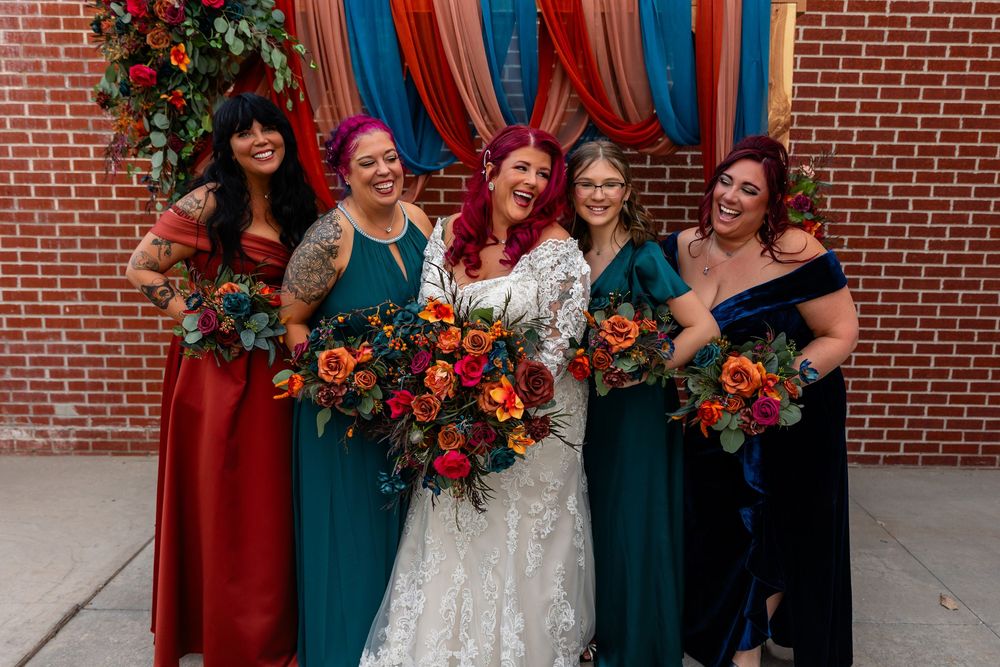 Bride in white lace gown laughs with four bridesmaids in teal and red dresses holding vibrant orange and red bouquets at Founders One Nine
