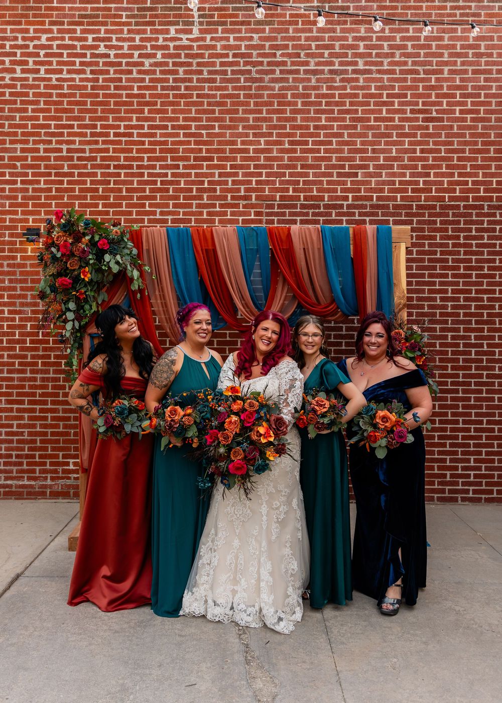 Bride and bridesmaids in teal and red gowns holding floral bouquets pose before a brick wall with colorful draping at Founders One Nine
