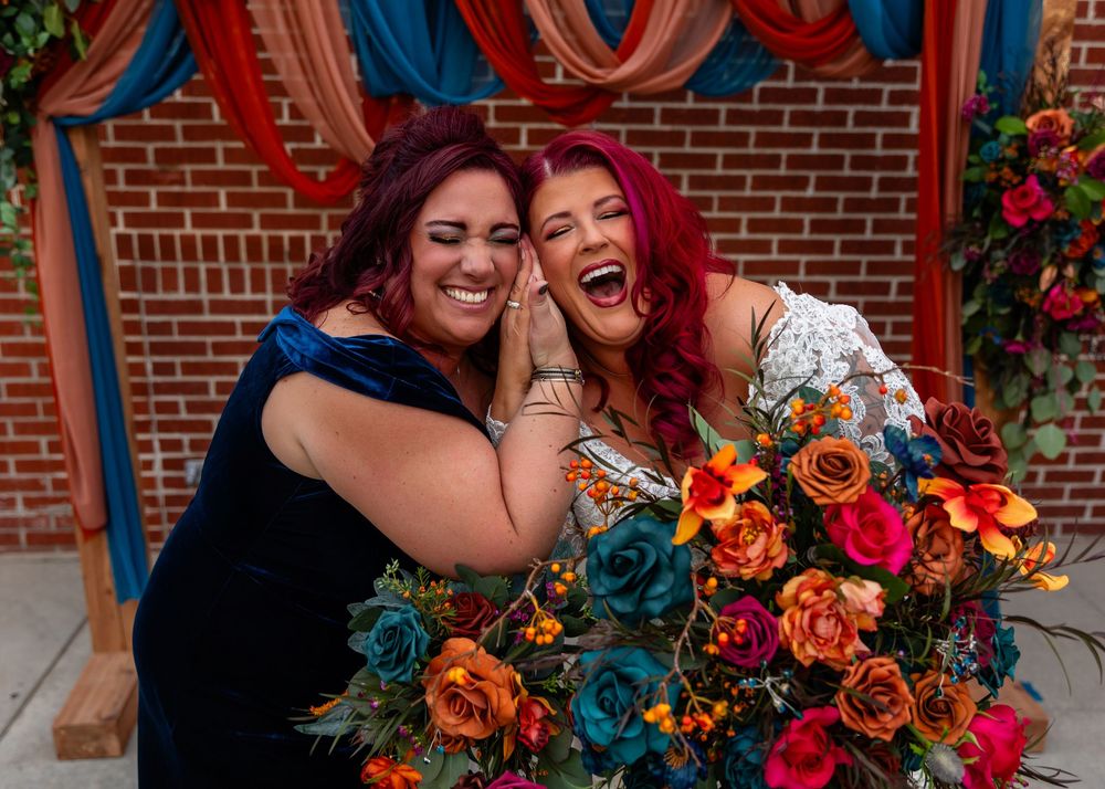 Two joyful women with red hair celebrate with vibrant orange and teal floral bouquets at a colorful wedding at Founders One Nine in Omaha