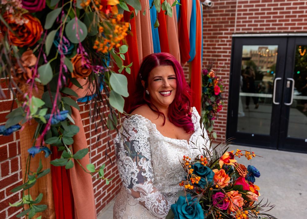 Radiant bride with red hair in lace wedding gown holds colorful bouquet beside vibrant floral arch at Founders One Nine in Omaha, NE.