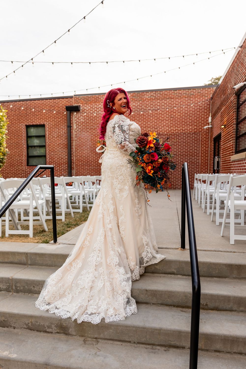 Bride in lace wedding gown with red hair holds colorful bouquet on outdoor stairs at brick venue at Founders One Nine in Omaha, NE.