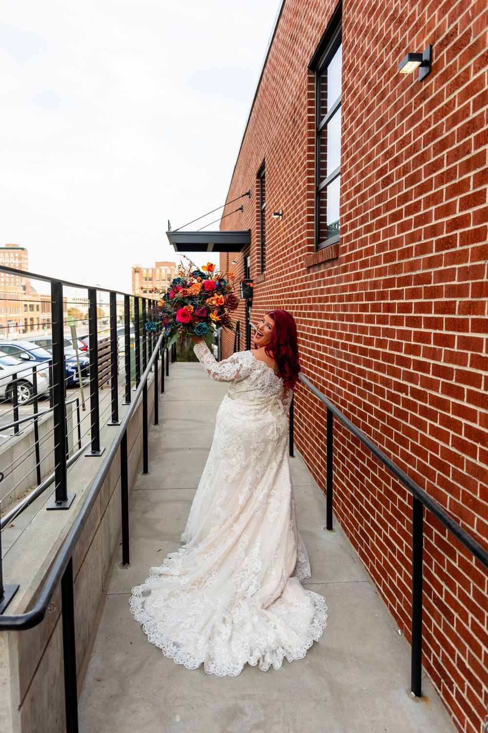 Red-haired bride in lace wedding gown holding colorful bouquet on brick building balcony with city view at Founders One Nine in Omaha, NE.