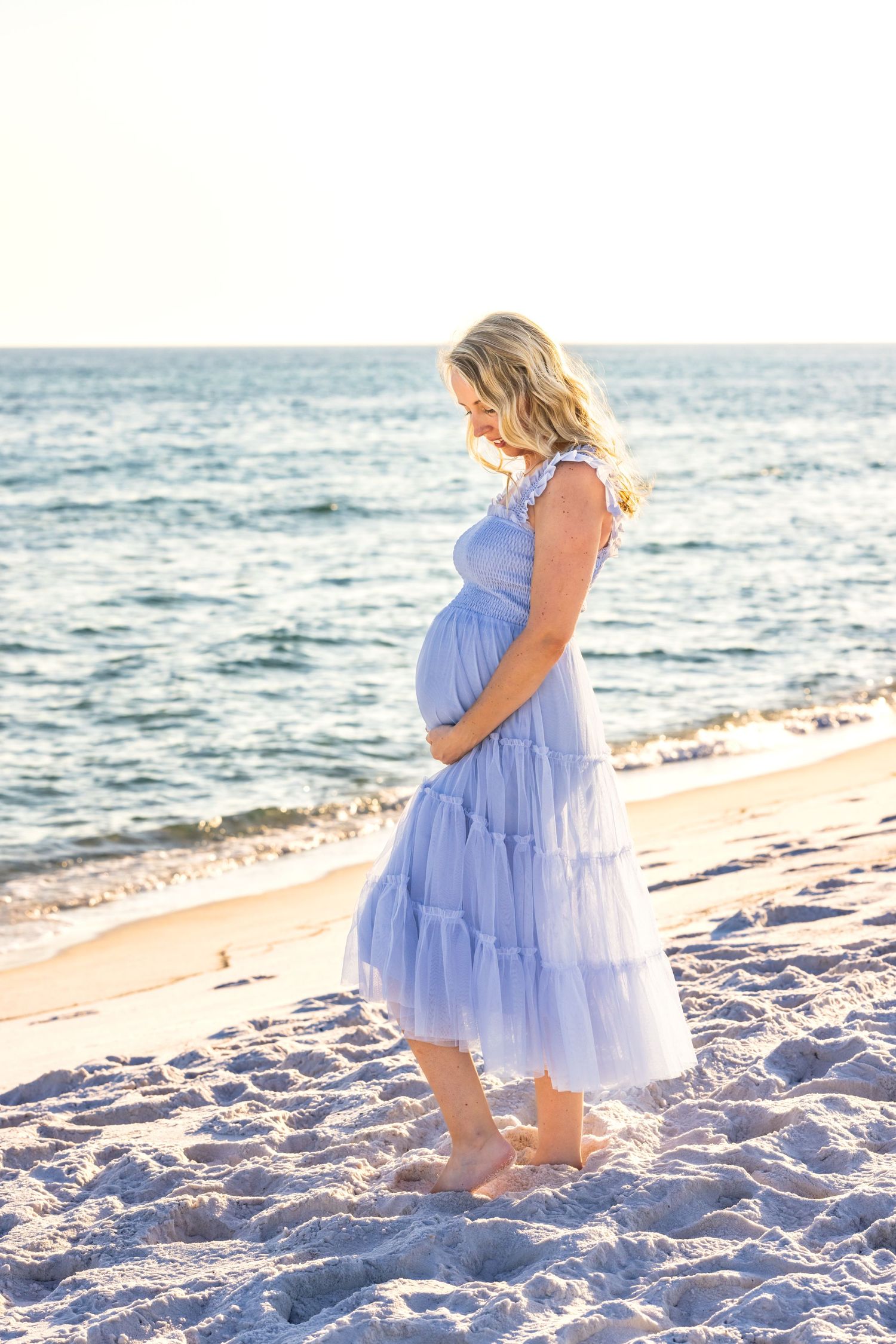 Woman in flowing periwinkle dress stands on beach during maternity photoshoot.