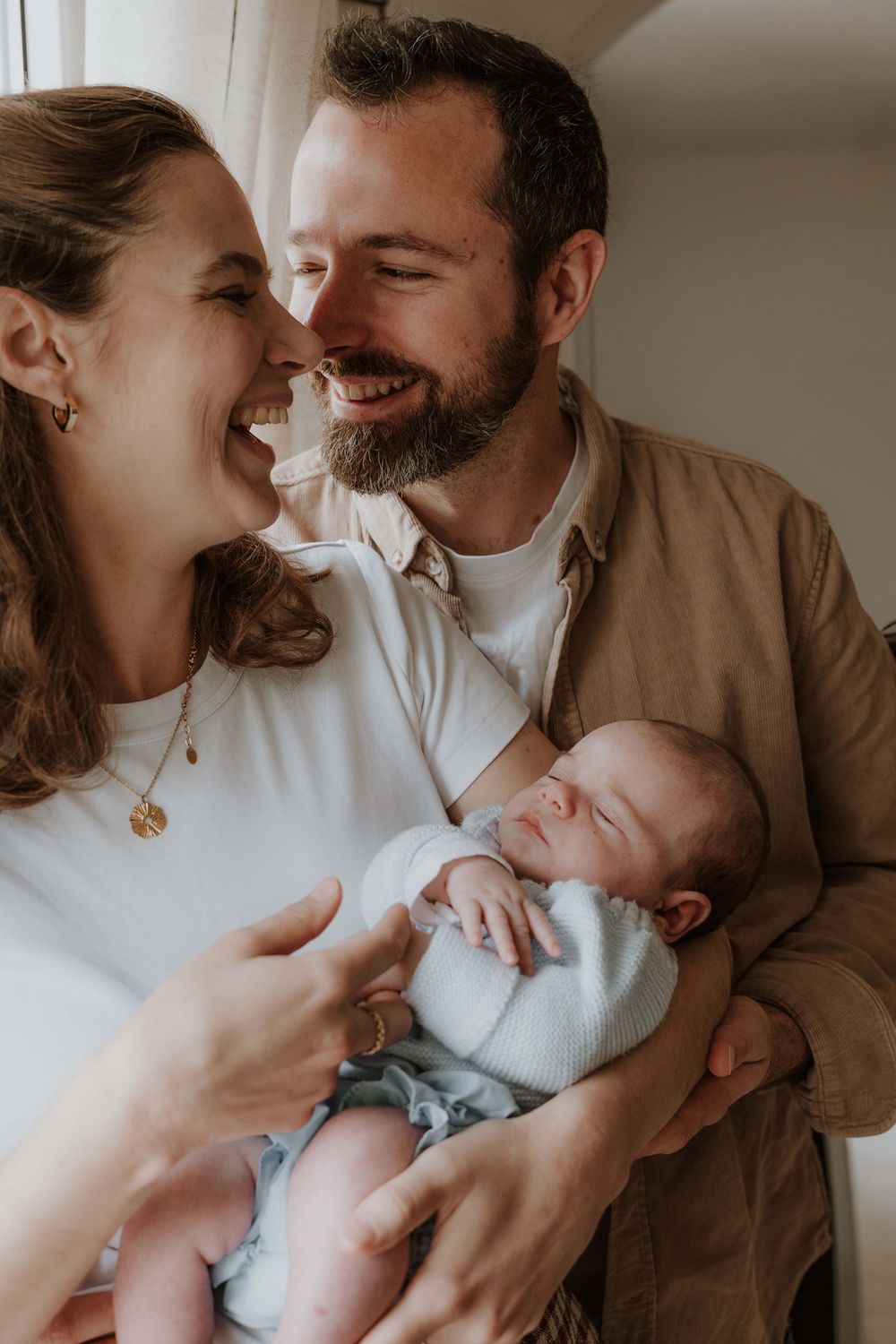Happy couple smiling at each other while gently cradling their sleeping newborn baby in a bright, cozy room.