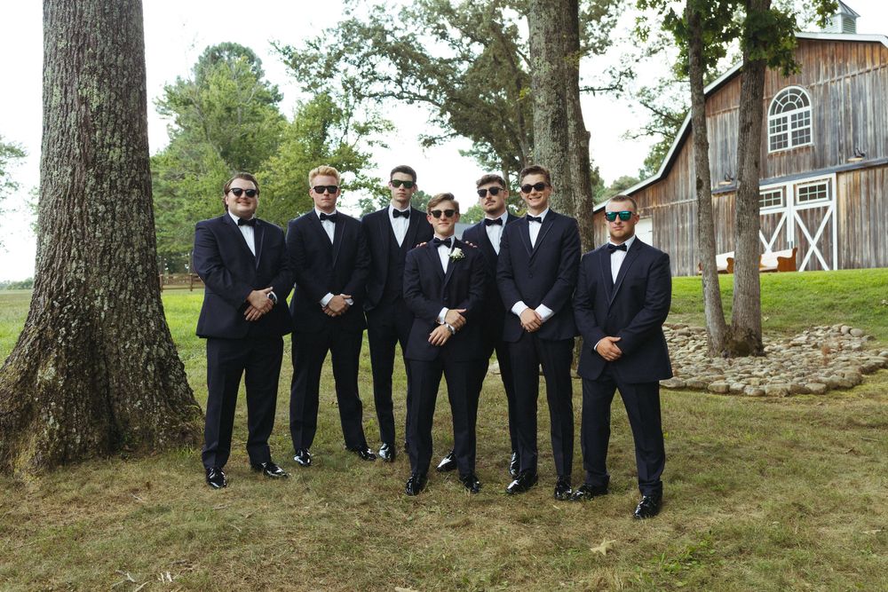 Groomsmen in black tuxedos pose together under large trees near brick building.