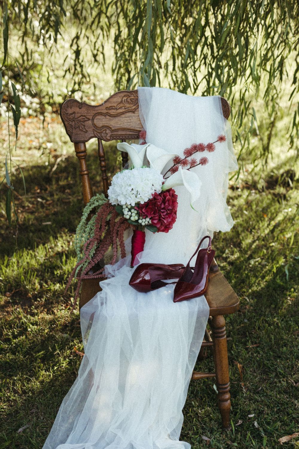 White wedding dress with red floral accents draped over vintage wooden chair outdoors.