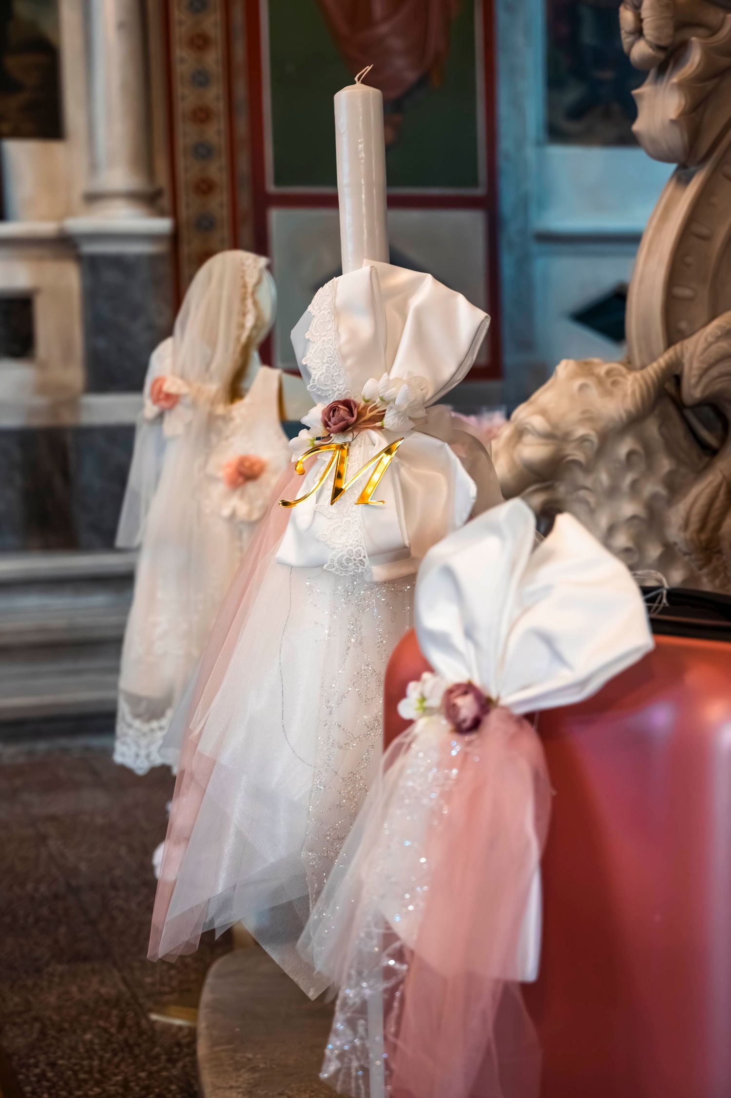 Decorative white baptismal candles adorned with tulle and ribbons displayed in a religious setting.