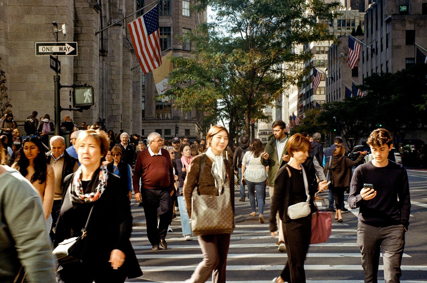 Crowds of pedestrians cross a busy intersection in Manhattan with American flags visible.