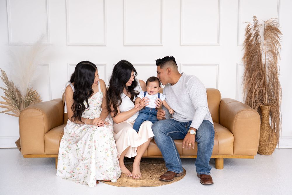 Family of four with parents and baby sitting on a tan couch in a bright white studio setting.