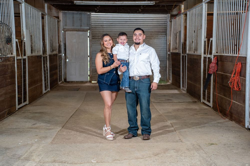 Family of three posing in a horse stable barn aisle, with stall doors on both sides.