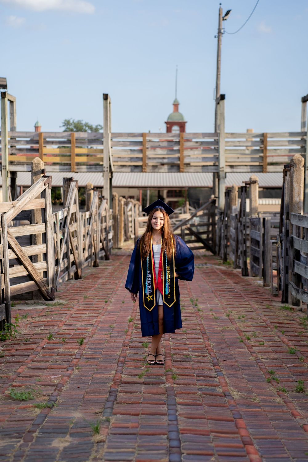 Graduate in navy cap and gown walks brick path flanked by wooden fences, smiling outdoors on a sunny day.