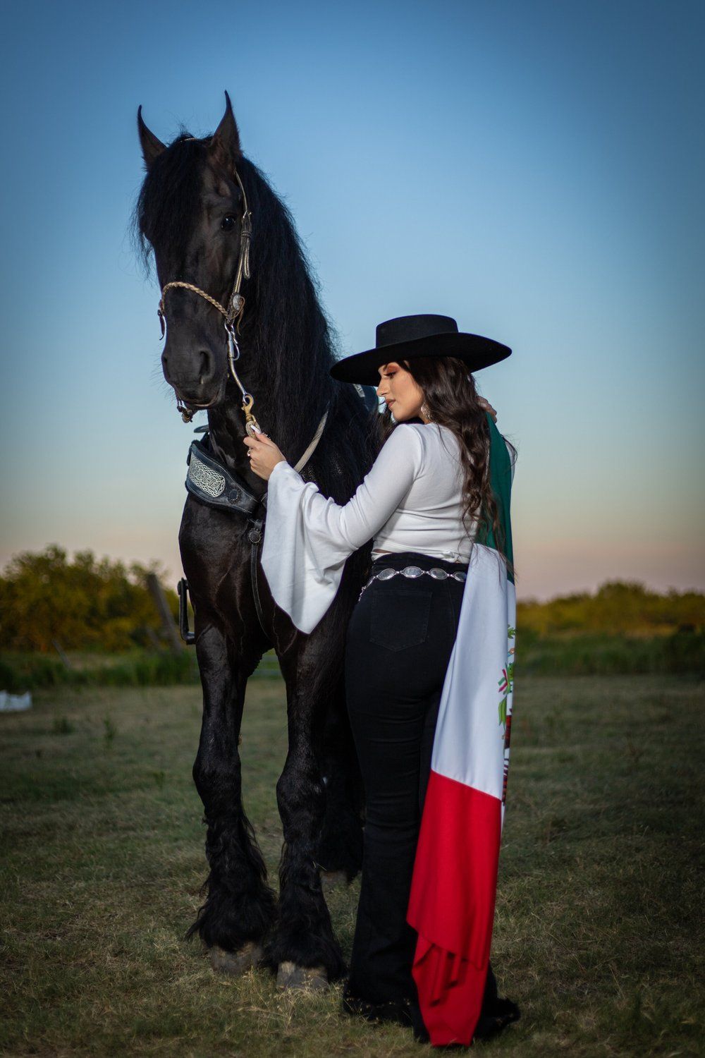 Person in traditional riding attire standing with a black horse in an open field at sunset.