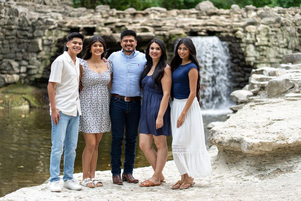 Family of five posing together outdoors near a scenic waterfall and stone landscape on a sunny day.