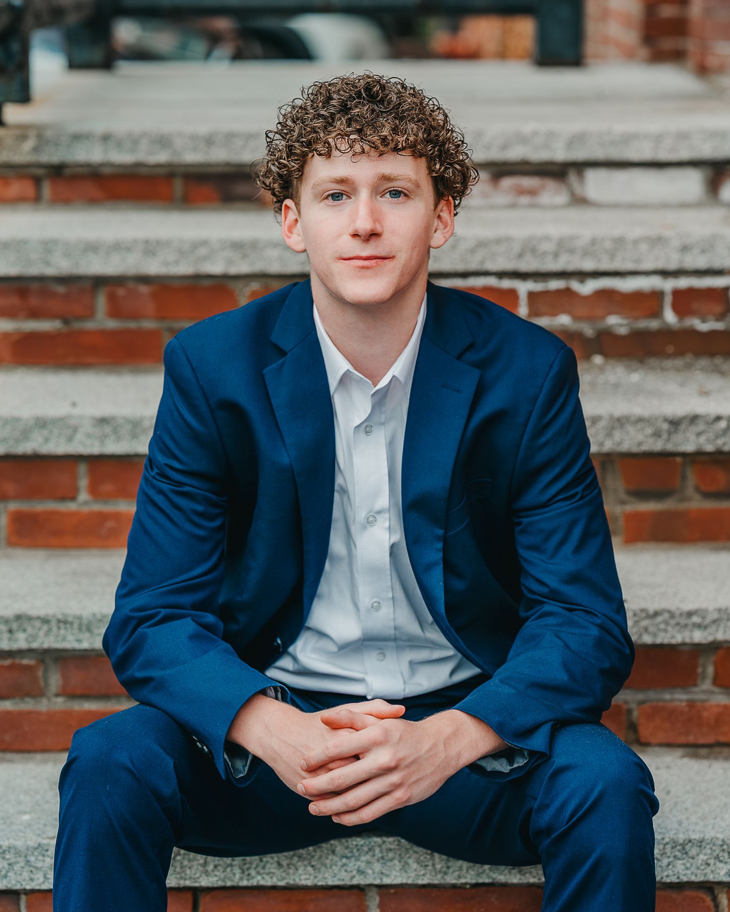 A senior boy in downtown Manchester, NH wearing a navy blue suit and white shirt and posing for photos while seated on stone steps.