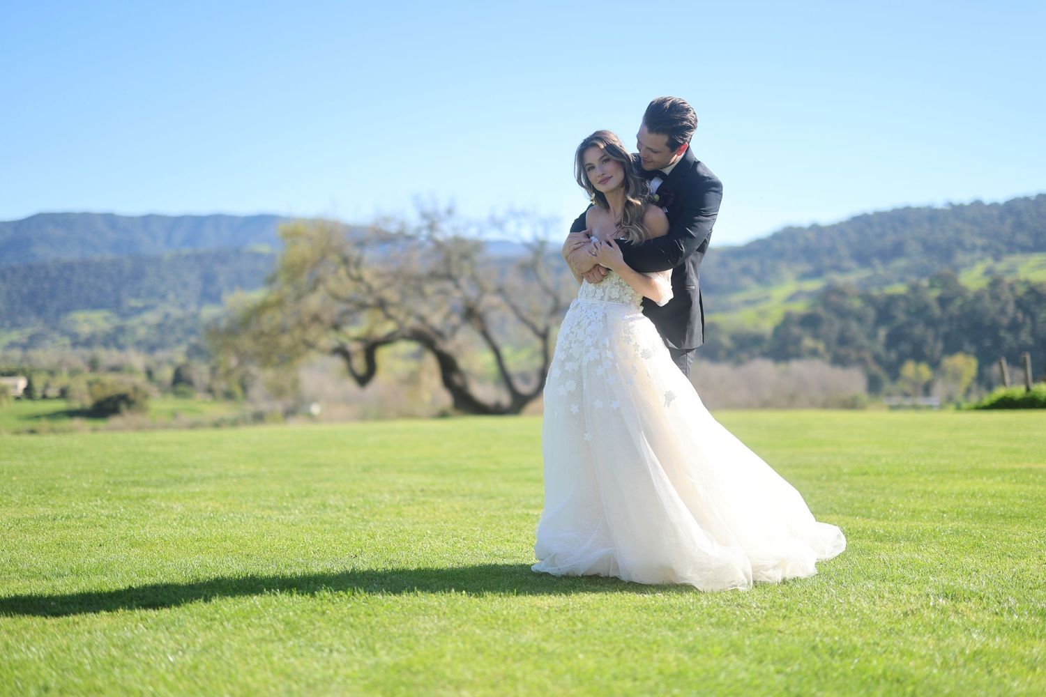 A romantic couple in a black outfit embrace while surrounded by lush greenery in a forest setting.