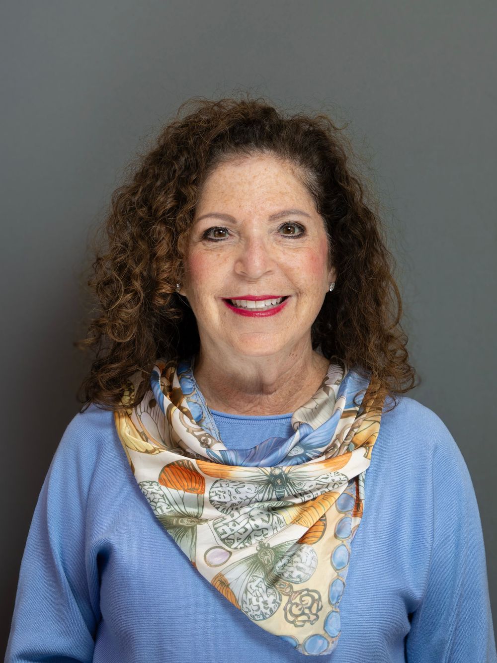 Smiling woman with curly brown hair wearing a blue sweater and floral scarf poses for a professional headshot.