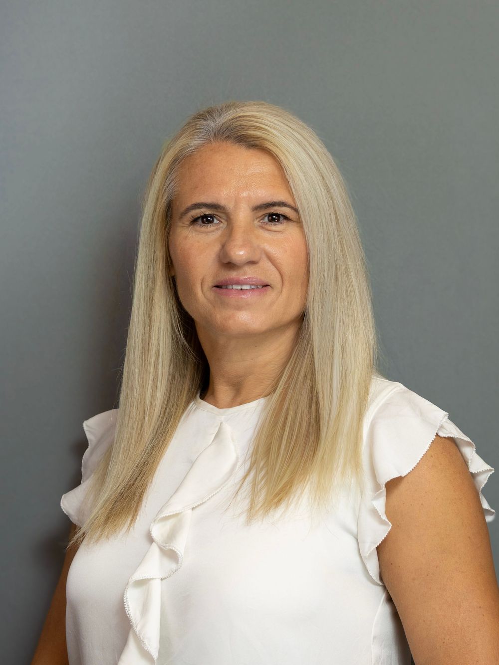Professional headshot of a blonde woman in a white ruffled sleeveless top against a gray background.