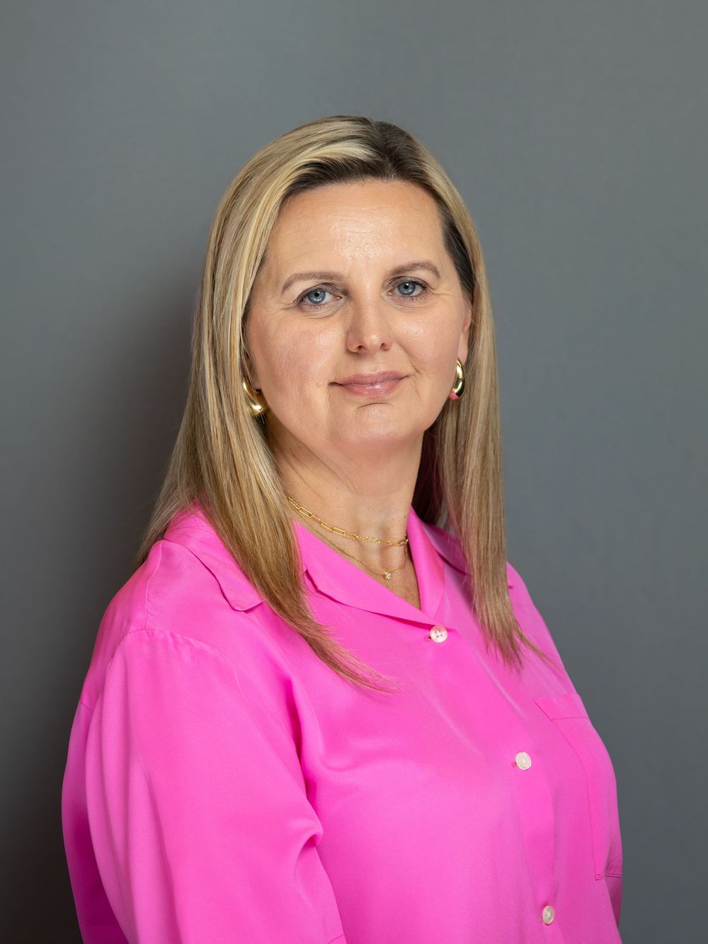 Professional headshot of a woman with blonde hair wearing a bright pink blouse, smiling against a gray background.