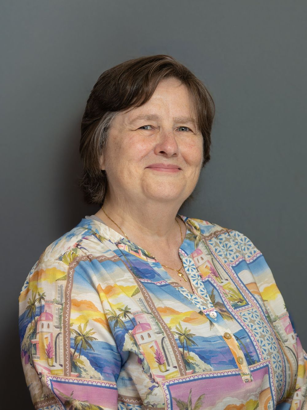 Smiling middle-aged woman with short brown hair wearing a colorful tropical print blouse, posed against a gray background.