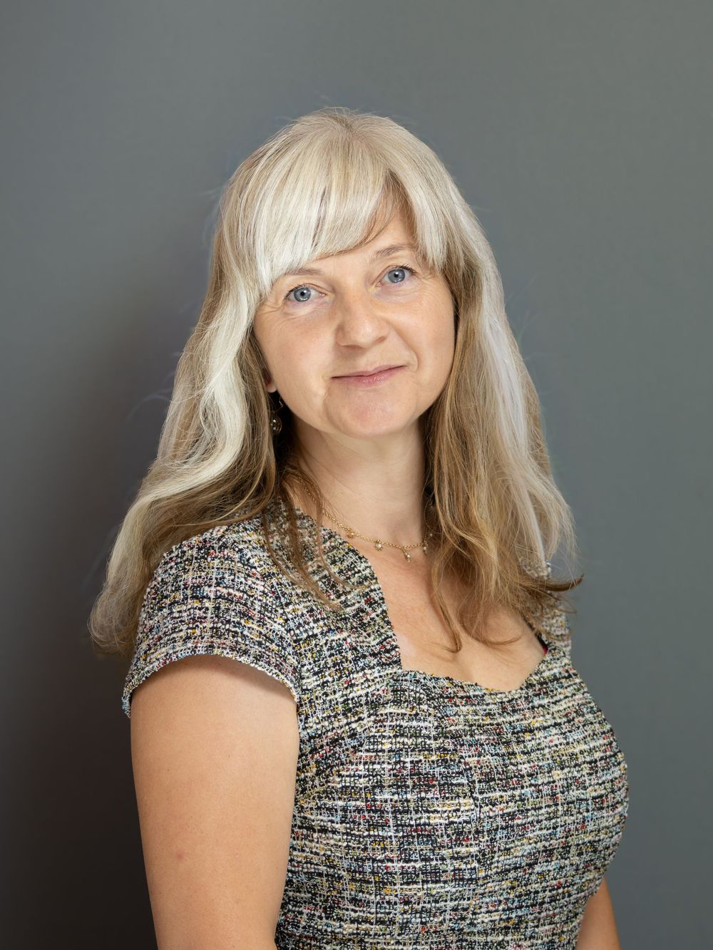 Professional headshot of a woman with long blonde-gray hair wearing a tweed top against a gray background.