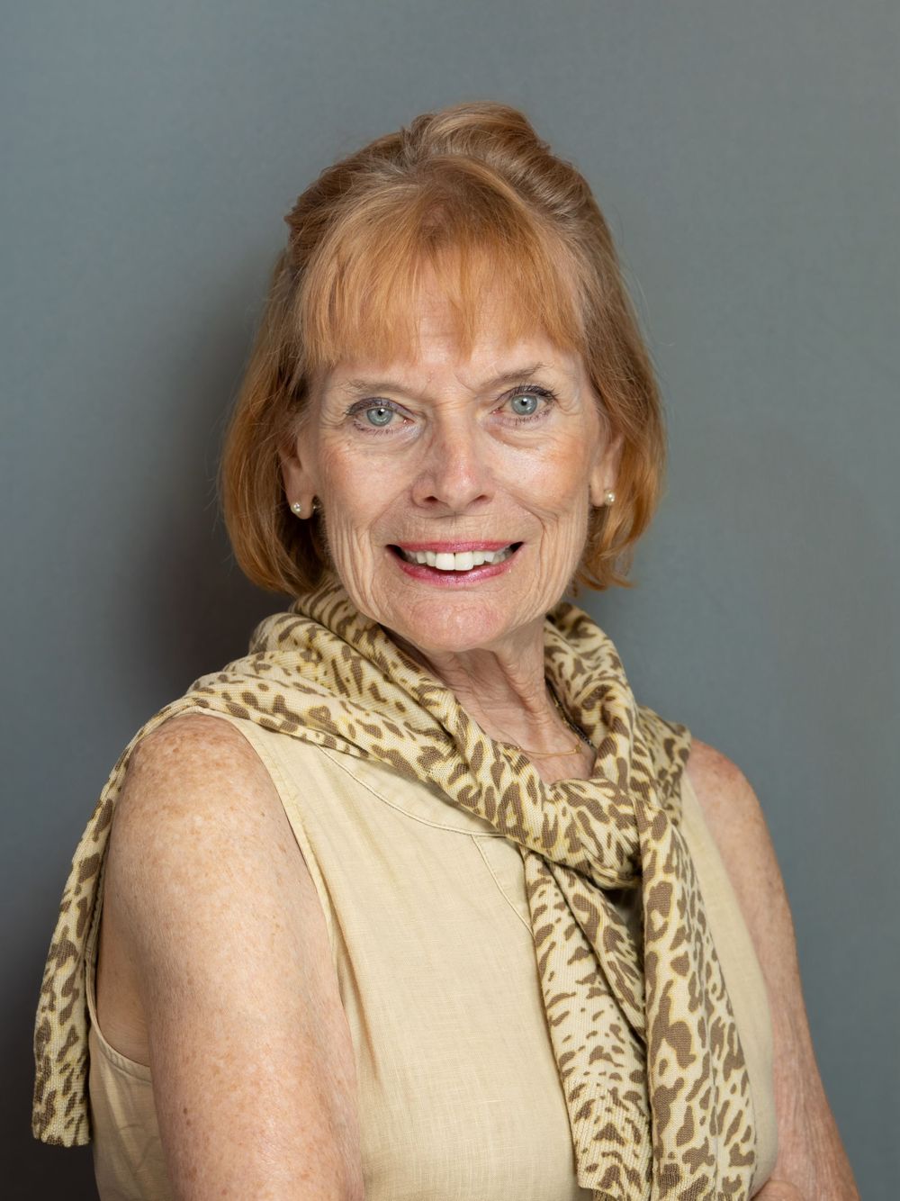 Smiling older woman with red hair wearing a tan sleeveless top and leopard print scarf against gray background.