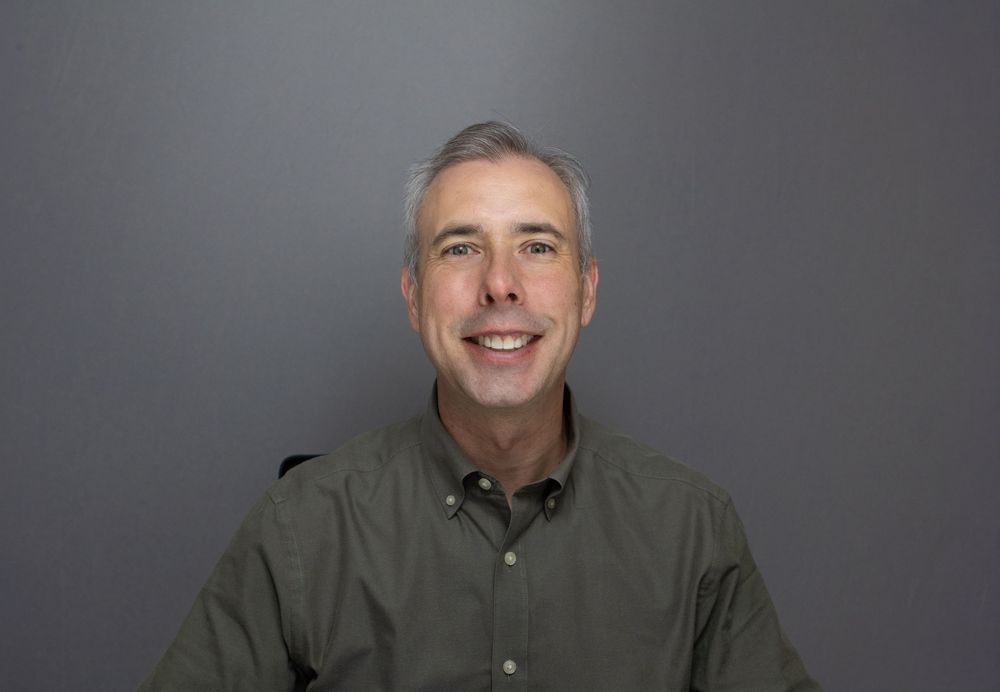 Smiling middle-aged man with gray hair wearing a olive green shirt against a gray background.