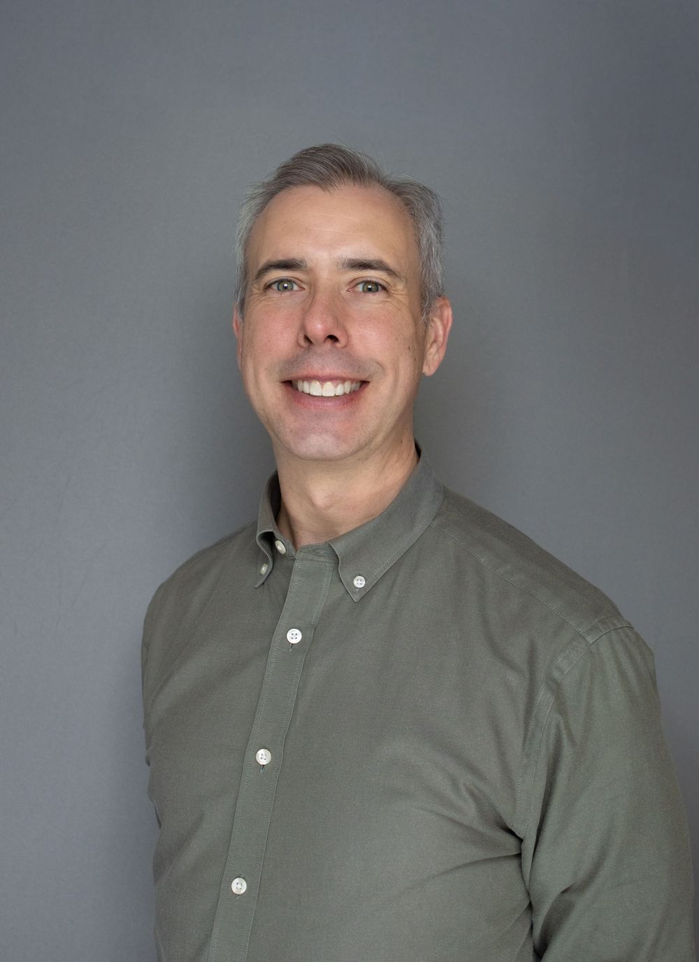Smiling middle-aged man with gray hair wearing an olive green button-up shirt against a gray background.