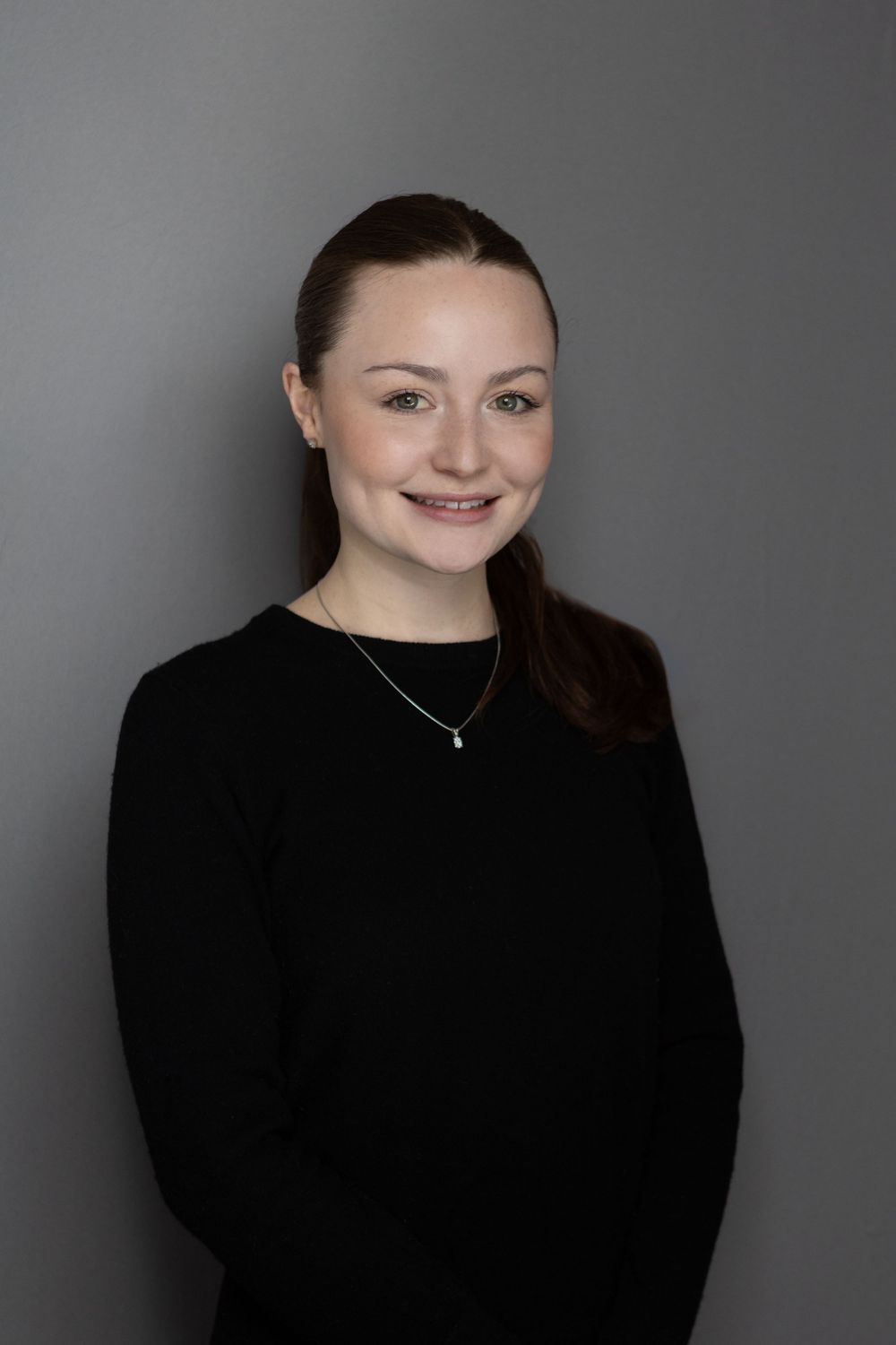 Young woman with dark hair smiling, wearing a black sweater and delicate necklace against a gray background.