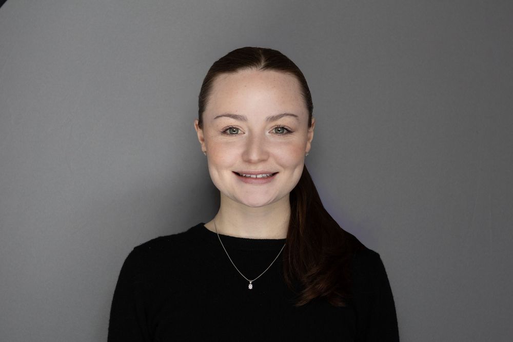 Young woman with brown hair smiling, wearing a black top and delicate necklace, against a gray background.