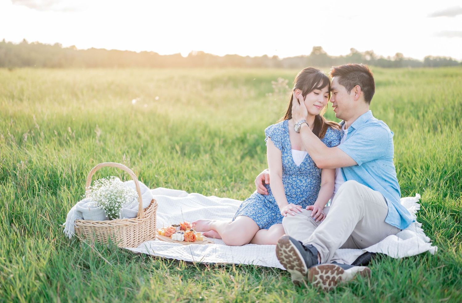 Two people enjoying a romantic summer picnic date in a grassy meadow with a woven basket at sunset.