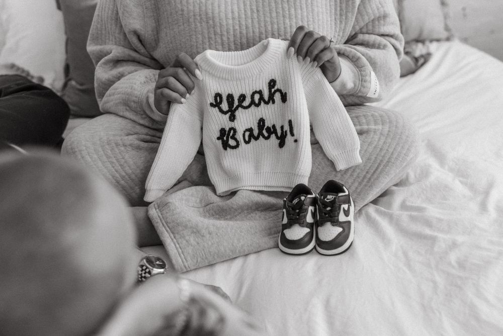 Black and white photo of a baby onesie with 'Yeah Baby' text and small sneakers arranged on cozy bedding.