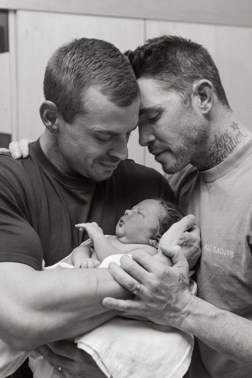 A tender black and white photograph shows two people sharing an intimate moment while holding a newborn baby.