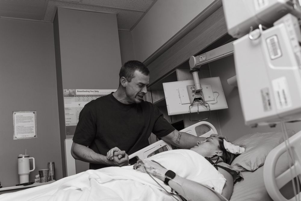 Black and white photo showing a caregiver in scrubs tending to a patient in a hospital room with medical equipment.