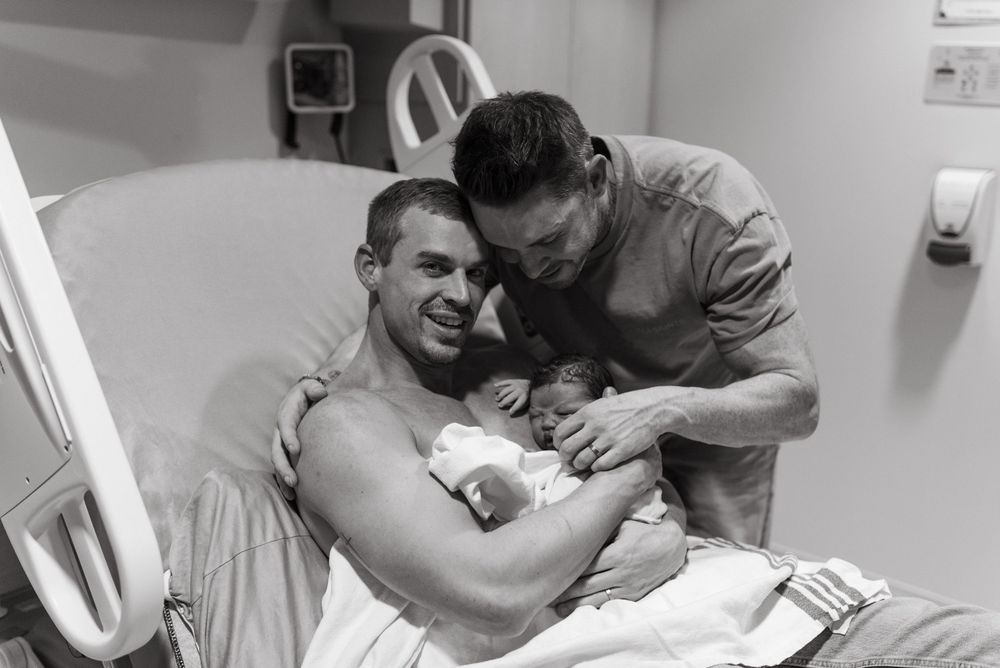 Black and white photo showing a proud new parent holding newborn baby in hospital room while another person leans in.