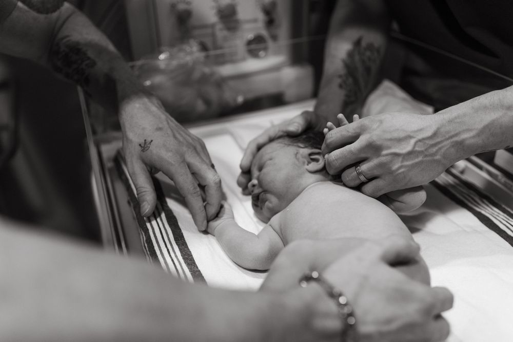 A newborn baby lies on a medical examination scale while medical staff hands perform measurements in black and white.