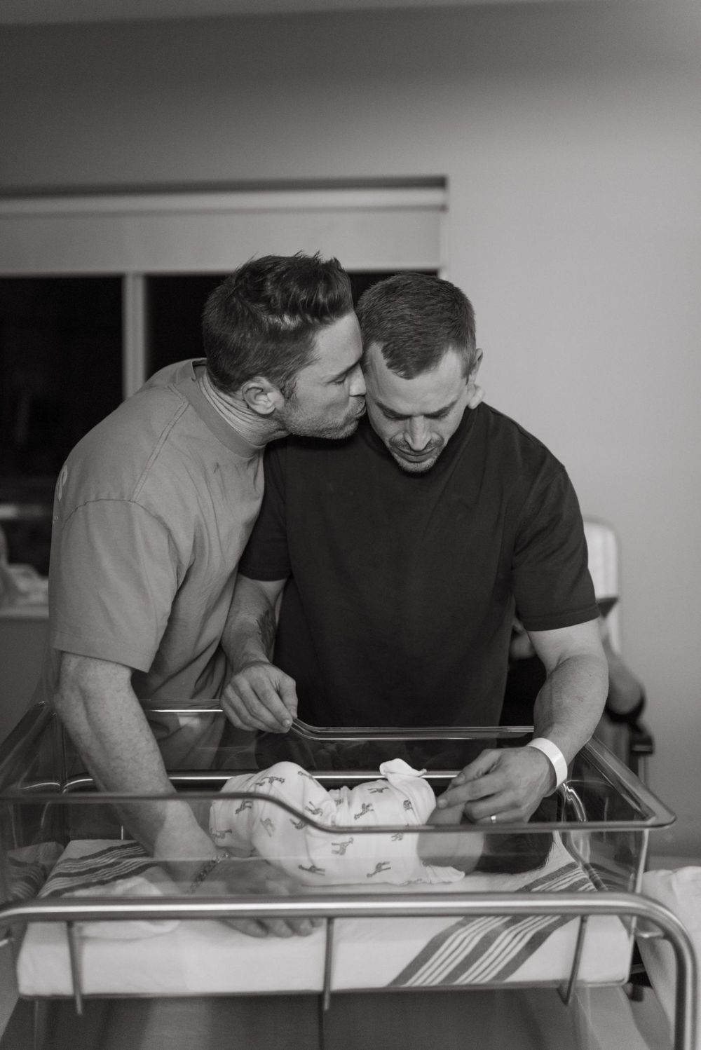 Two people lean over and look into a hospital bassinet in a black and white photo series.