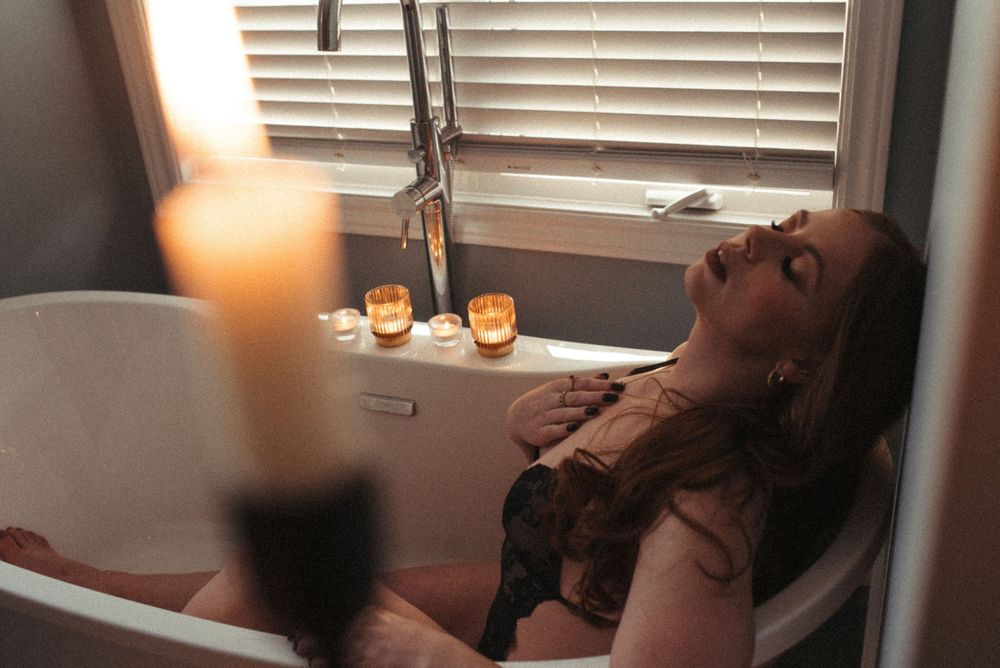 Dimly lit bathroom with candles glowing near a soaking bathtub by a window with venetian blinds.