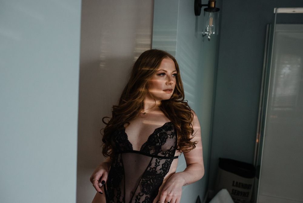 Model in a lacy lingerie setpiece gazes seductively at the camera in a dark and moody boudoir photoshoot setup.