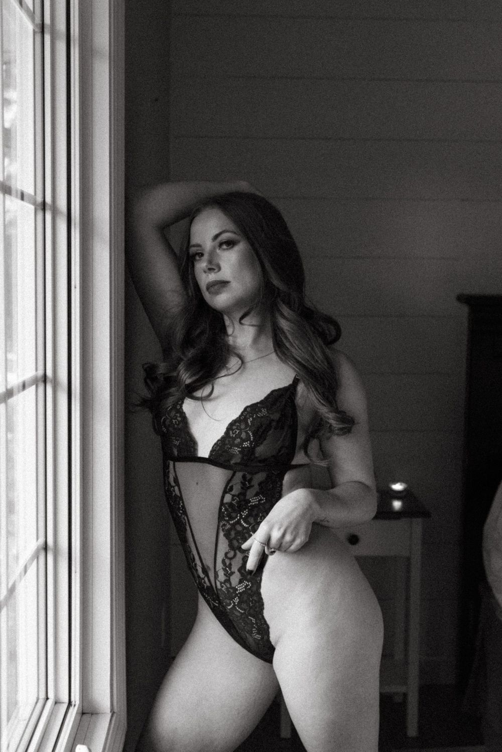 Black and white intimate portrait of a person in lace bodysuit posing by a window.