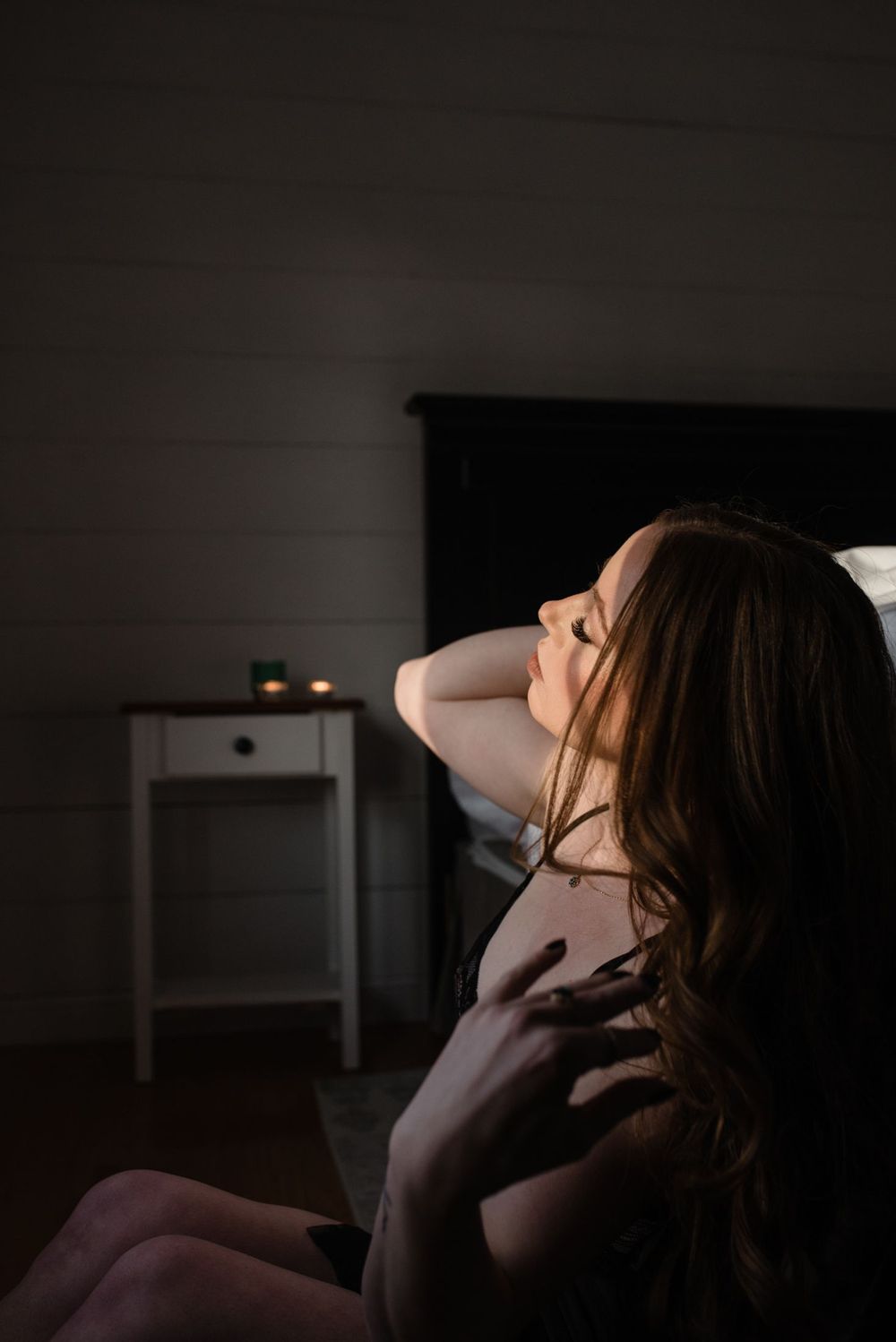 A sensual boudoir portrait in moody lighting shows a woman with flowing hair in an intimate bedroom setting.