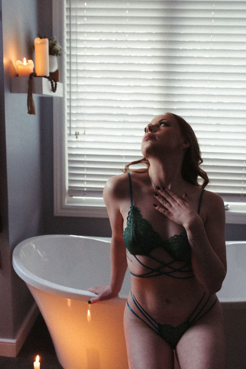 Moody boudoir scene with candlelight and white bathtub against venetian blinds in intimate setting.