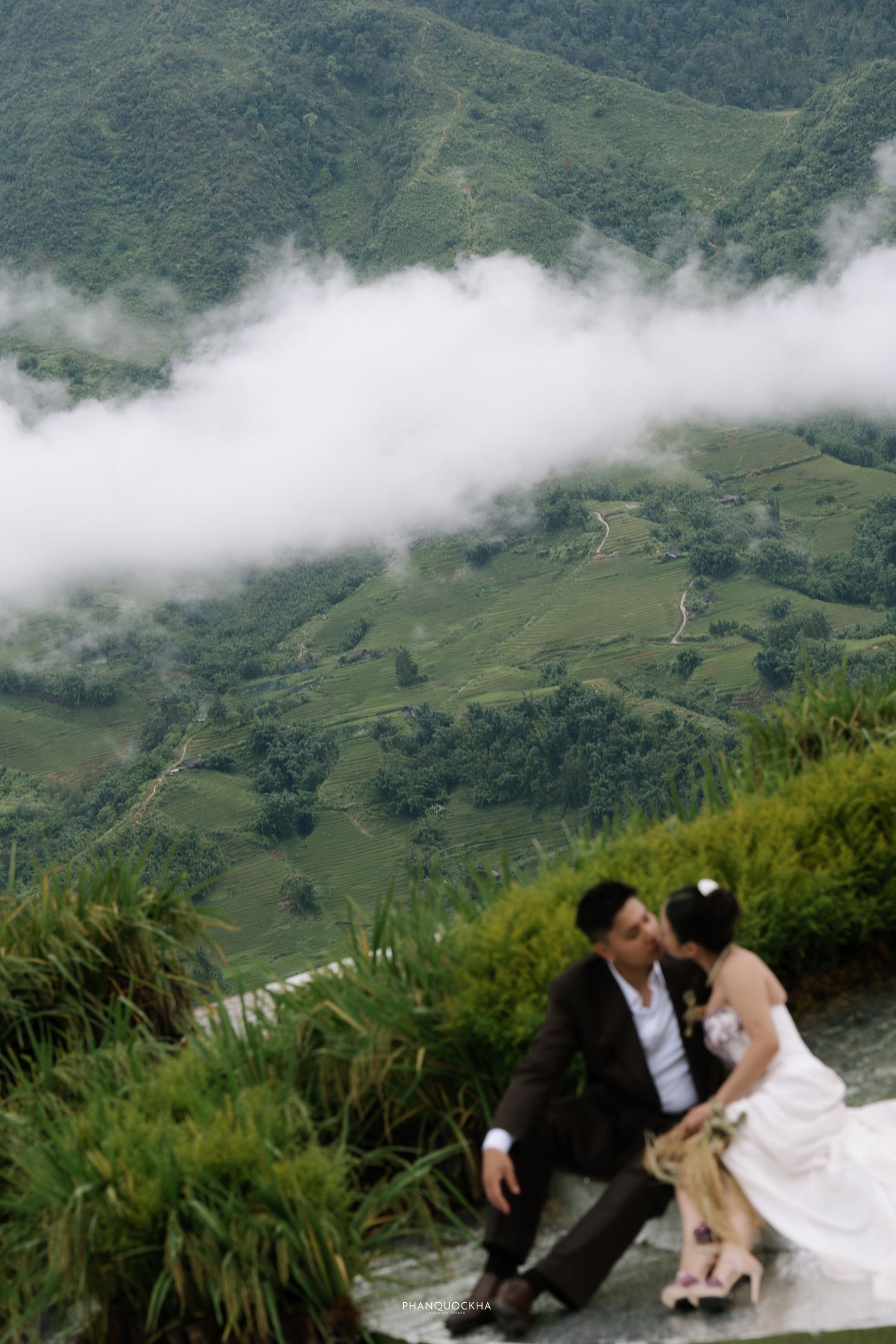 Wedding couple shares a kiss on hillside terrace with dramatic cloudy green mountain valley below.