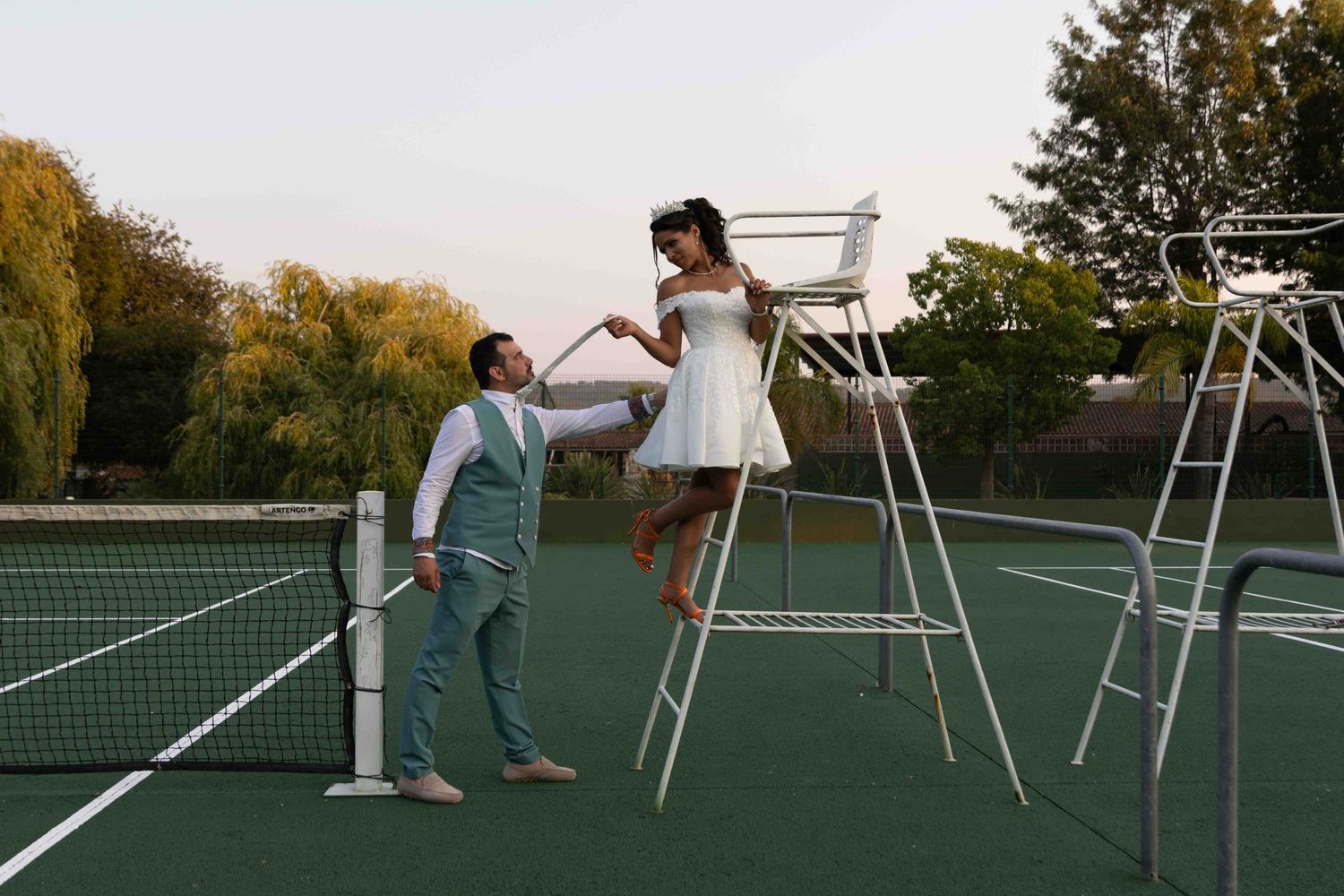 Creative wedding photo on tennis court with couple posing on umpire chairs.
