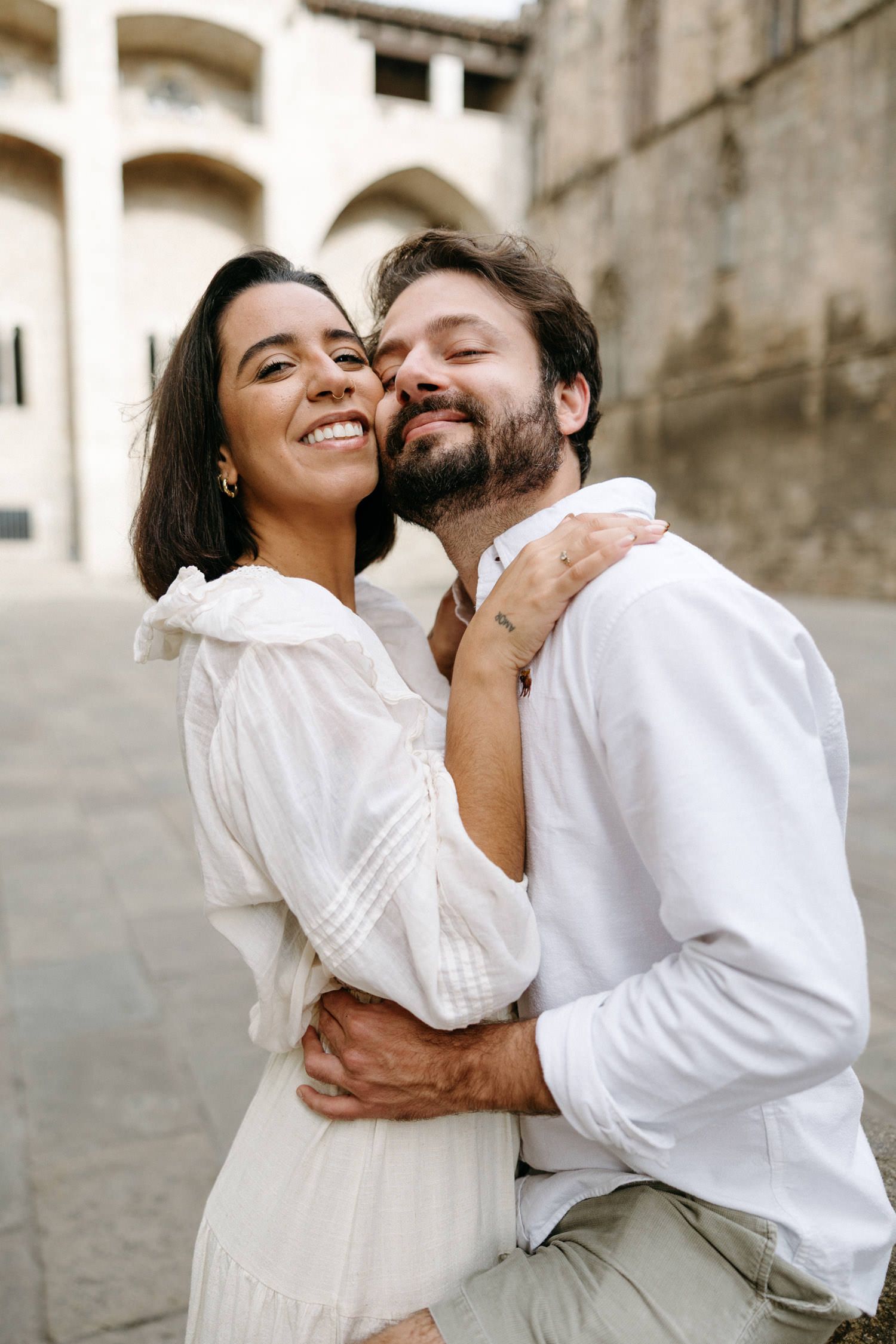 A loving couple in matching white outfits embracing against a historic stone building backdrop.