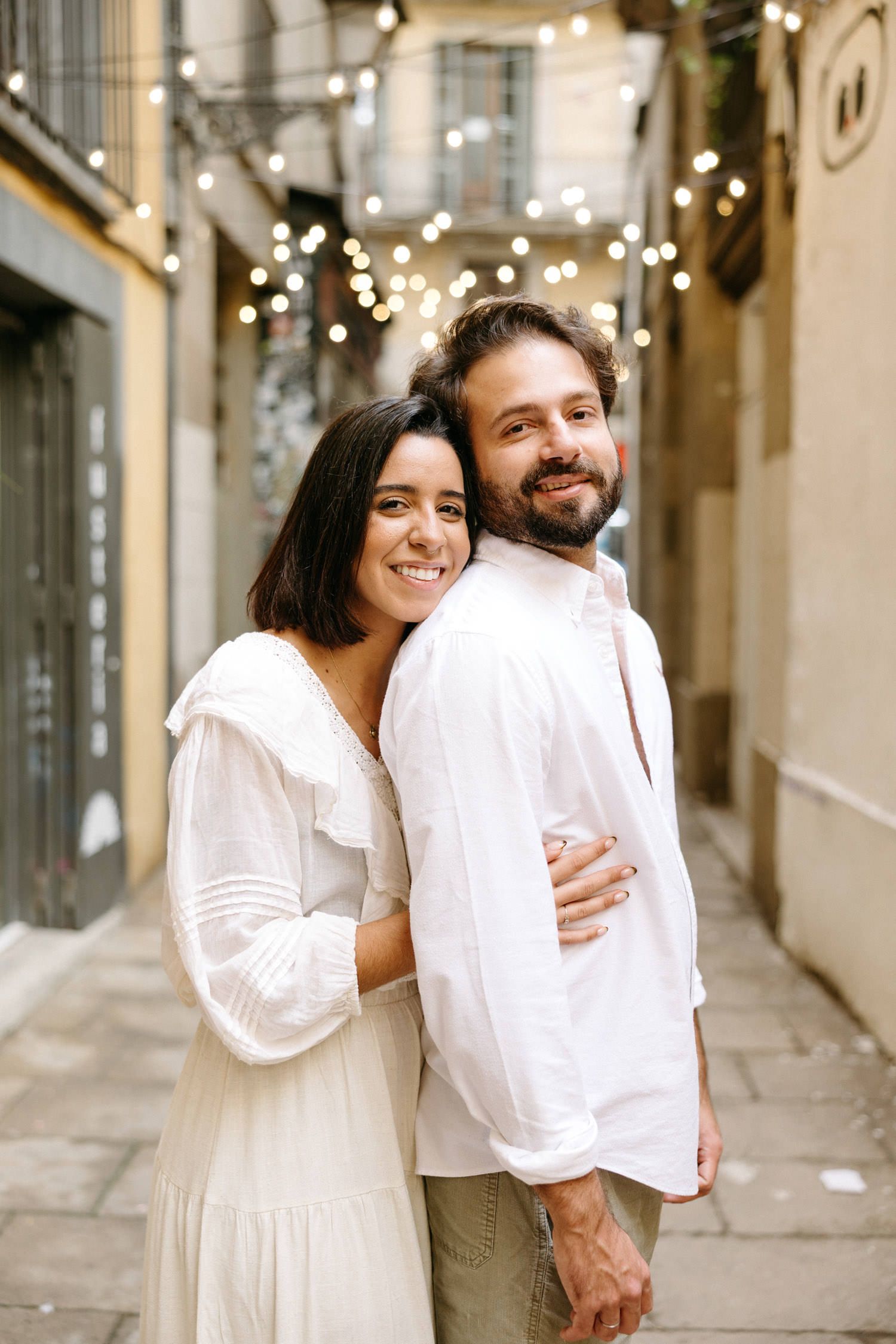 A romantic couple in white outfits embracing in a charming European alleyway with twinkling lights overhead.