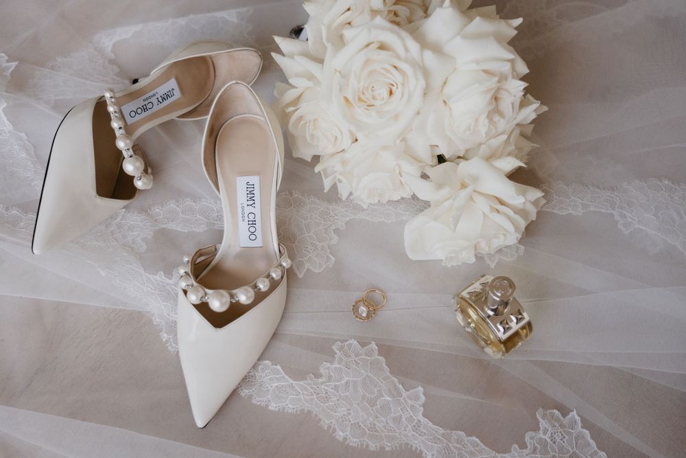 White wedding accessories including heels, pearls, lace veil, roses and jewelry arranged on a tulle background.
