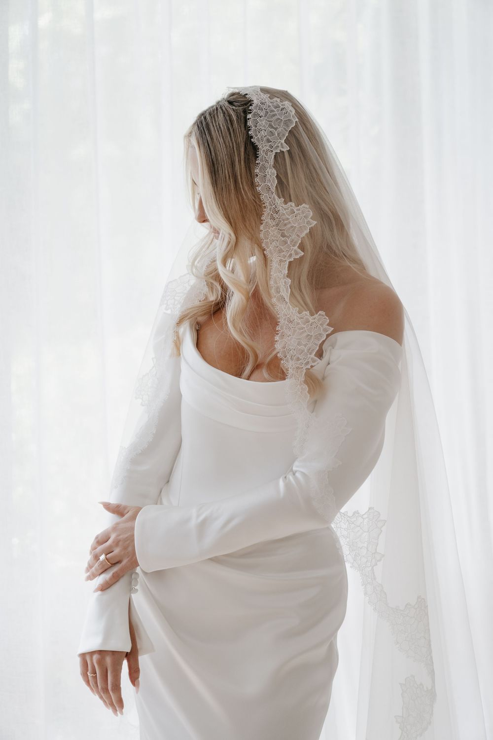 Woman wearing elegant white bridal robe with delicate lace details and billowing sleeves poses against white backdrop.